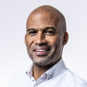 Carlton Williams clasping hands and smiling wearing a white shirt and blue jeans on a white background