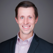 Austin Arnold smiling wearing a checkered shirt and suit jacket on a gray background.