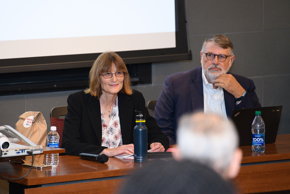 Two panelists, female on the left, male on the right in front of a white screen. Blurry back of two attendees heads in the foreground.