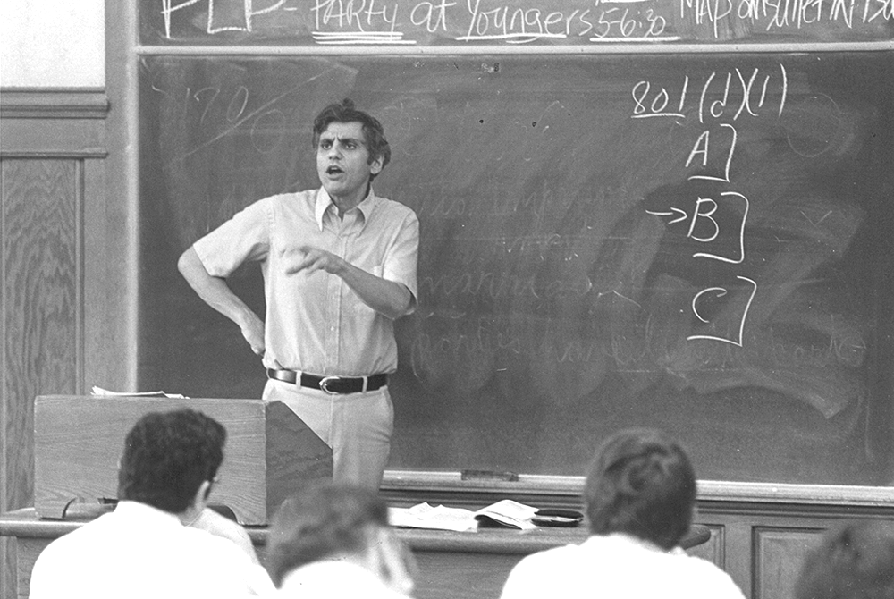 photo of Rossi giving a lecture in a classroom, standing in front of a chalkboard, talking