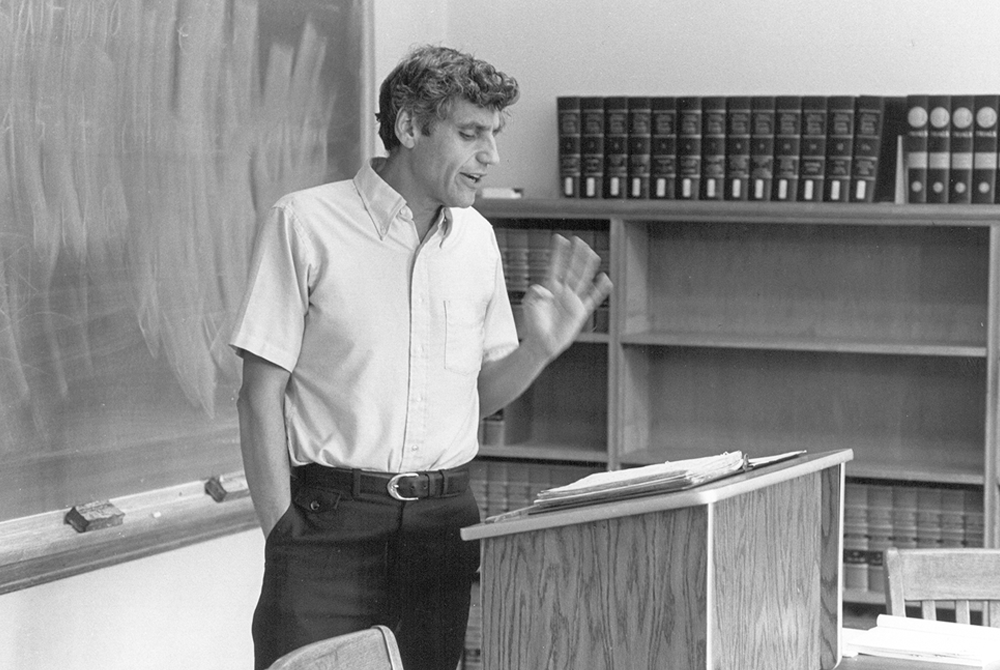 photo of Rossi giving a lecture in a classroom, standing in front of a chalkboard, talking- there is a podium in front of him
