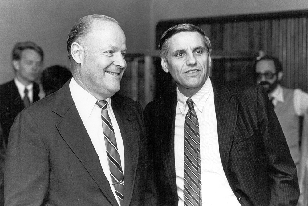 photo of Rossi standing next to another man talking at an event- both men are dressed in dark suits