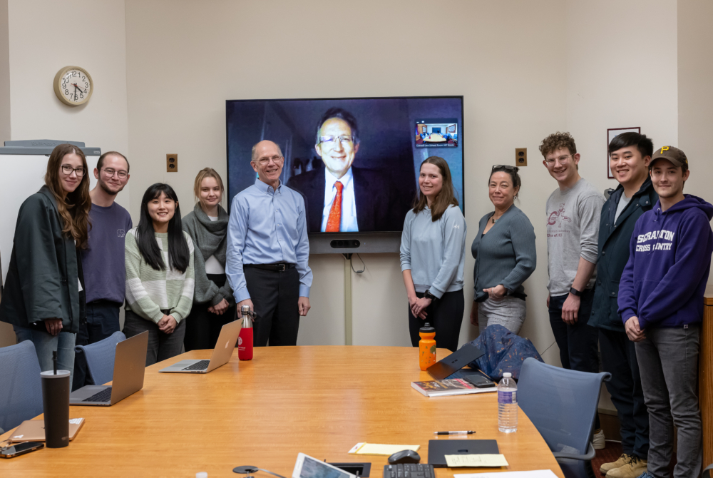 Group of students and faculty around a large screen with a guest speaker