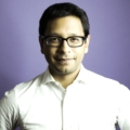 Headshot of a man wearing a white suit shirt and black frame glasses. He had dark hair and is standing in front of a purple background