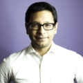 Headshot of a man wearing a white suit shirt and black frame glasses. He had dark hair and is standing in front of a purple background