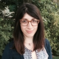 Afroditi Giovanopoulou outdoors wearing glasses and smiling with green leaves in background.